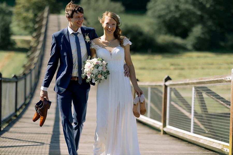 Das Brautpaar läuft auf einer Hängebrücke auf die Kamera zu, sie haben ihre Schuhe in der Hand..